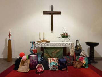 Blick auf den Altarraum der Heilandskirche mit Kreuz, Kerzen und Blumenschmuck. Vor dem Altar stehen zahlreiche Taschen verschiedener Art — darunter Schulrucksäcke, Handtaschen und eine Reisetasche — als Anschauungsobjekte für den Gottesdienst zum Gemeindefest.